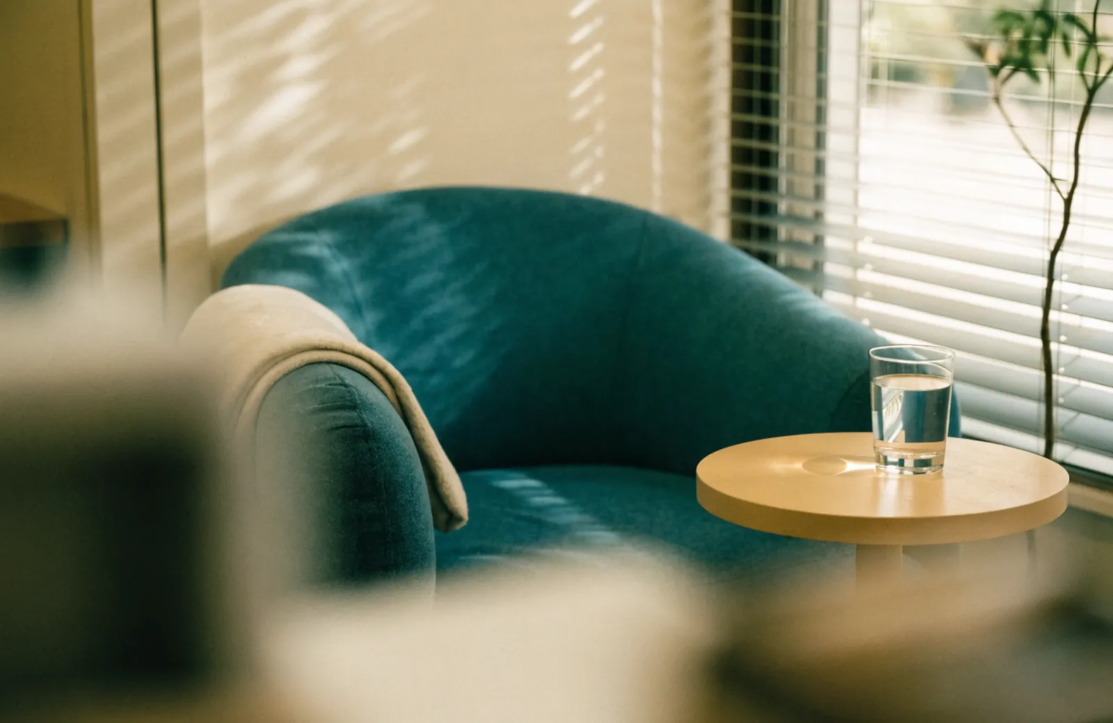 A quiet lounge with a soft armchair, a folded blanket and a glass of water by a window