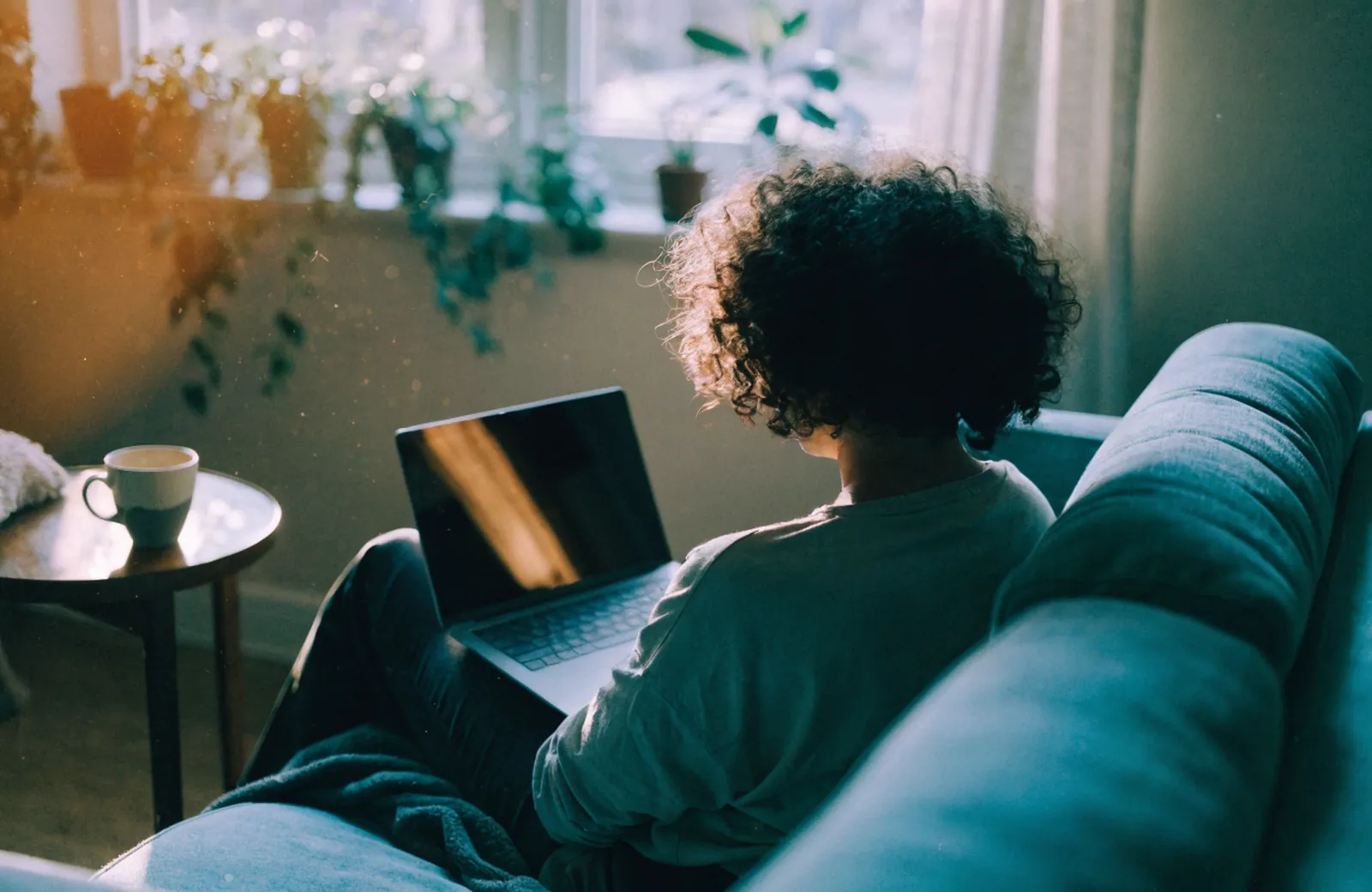 A person on a sofa at home with a laptop open for a video appointment, morning light through the window